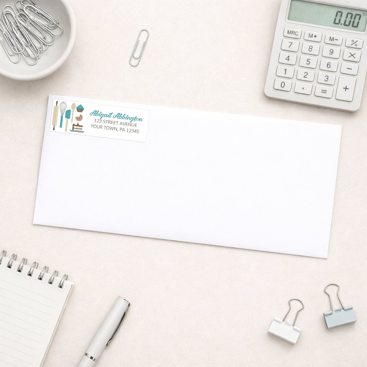 Turquoise Baking Address Label shown on a white envelope with everyday office supplies