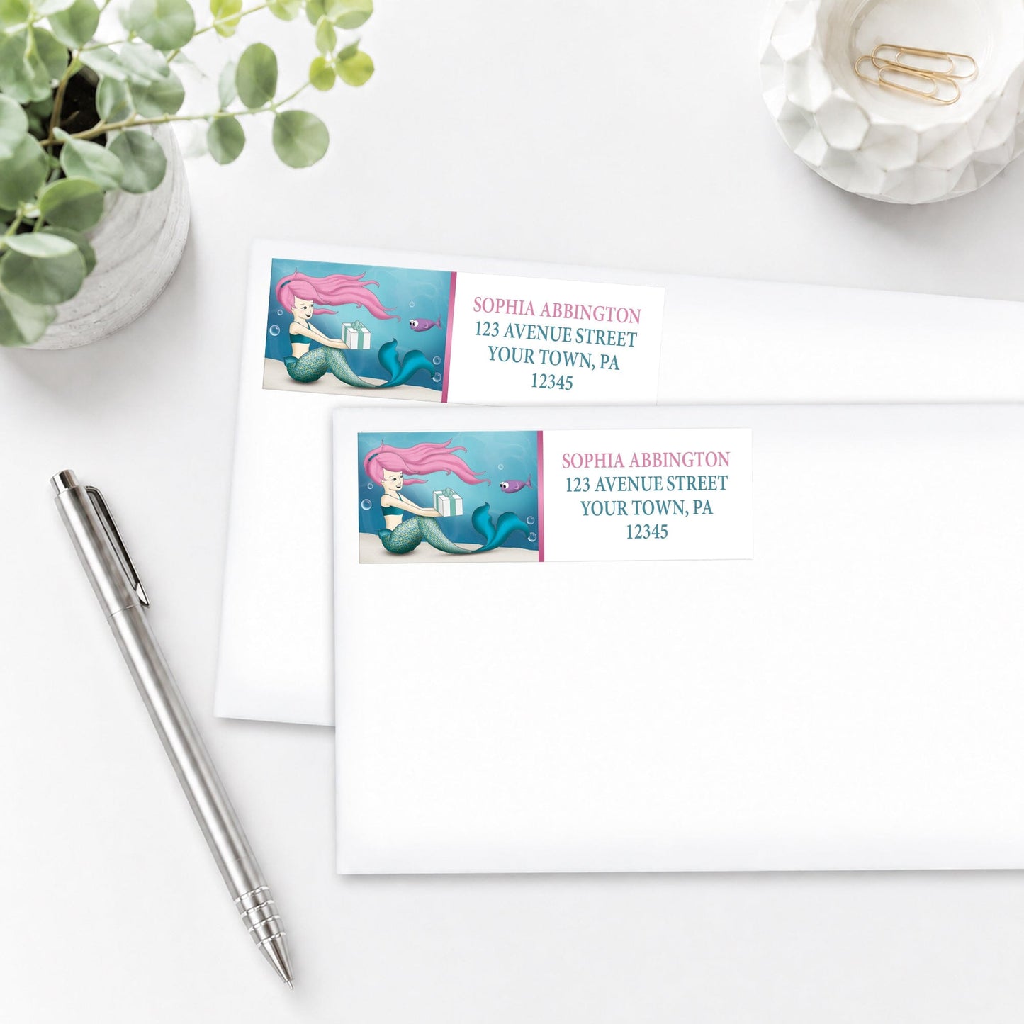 Mermaid-themed address labels on white envelopes with a pen and plant in the background.