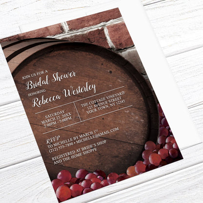 Bridal shower invitation with wooden barrel and grapes design, shown with a white envelope, on a wooden surface at an angle