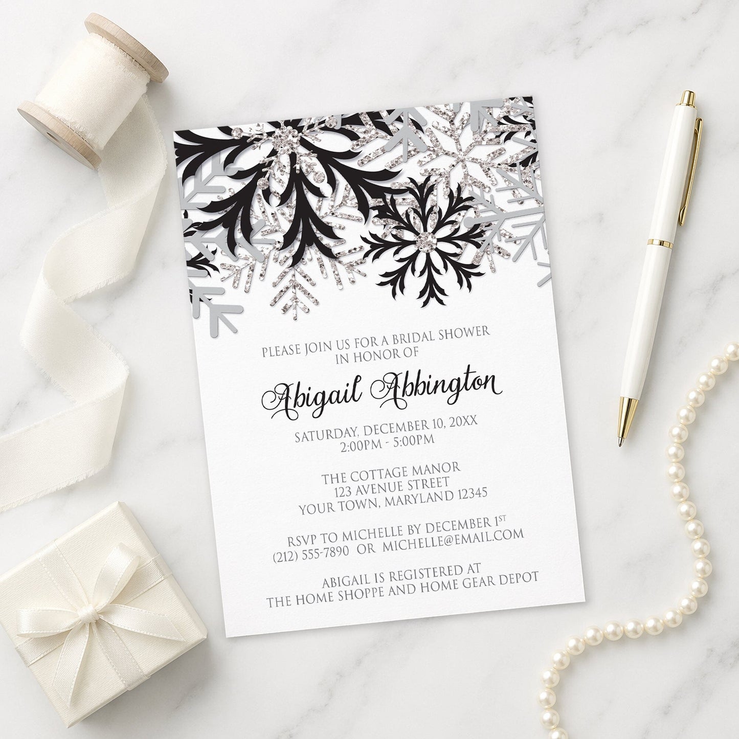 Winter Black Silver Snowflake Bridal Shower Invitation shown on a marble surface with a white gift box, pen, ribbon, and pearls.