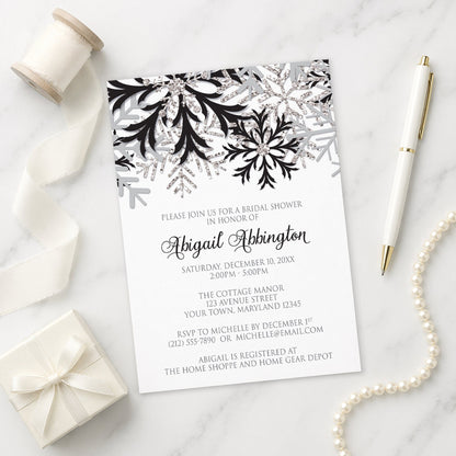 Winter Black Silver Snowflake Bridal Shower Invitation shown on a marble surface with a white gift box, pen, ribbon, and pearls.