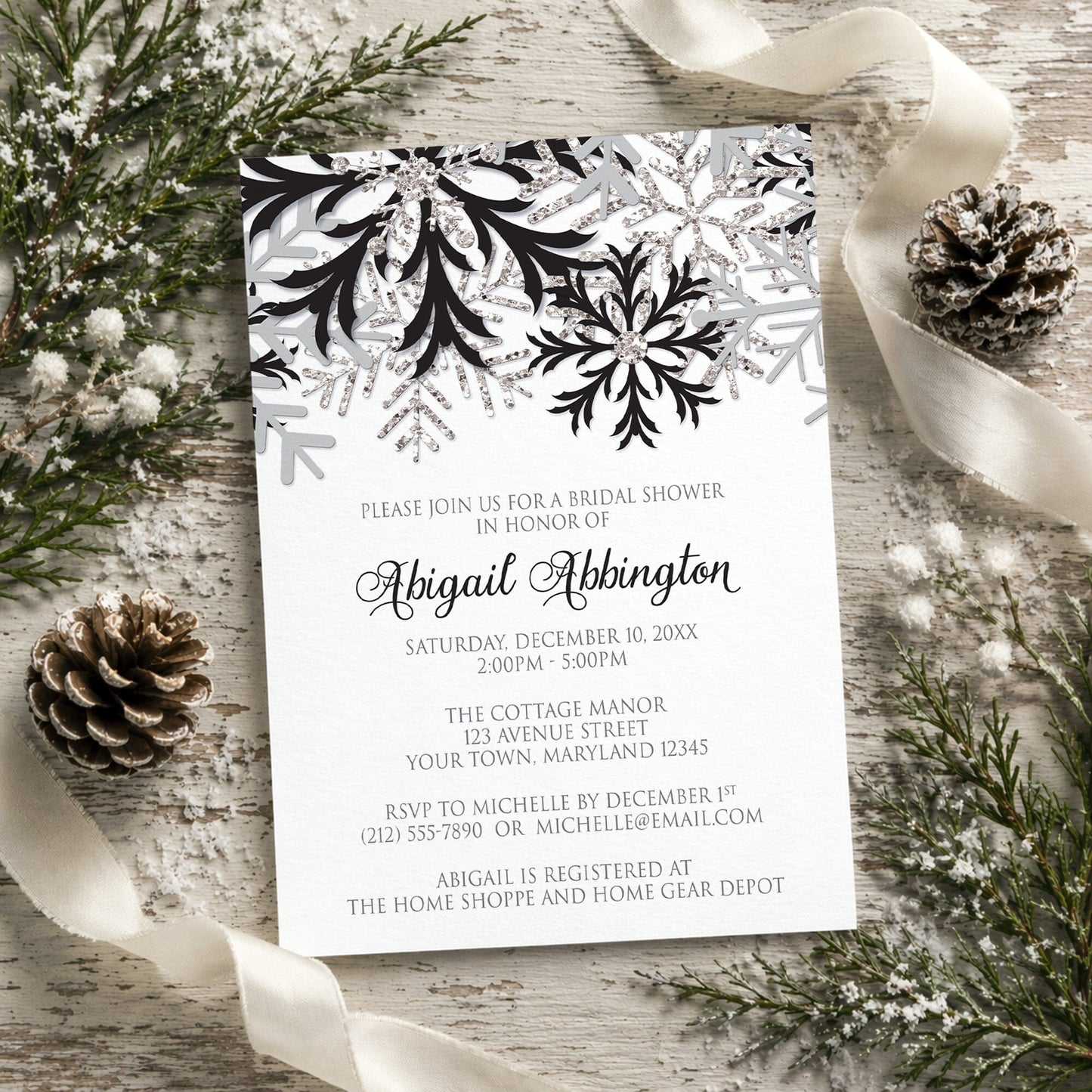 Winter Black Silver Snowflake Bridal Shower Invitation shown on a rustic surface with winter greenery, pine cones, and ribbons.