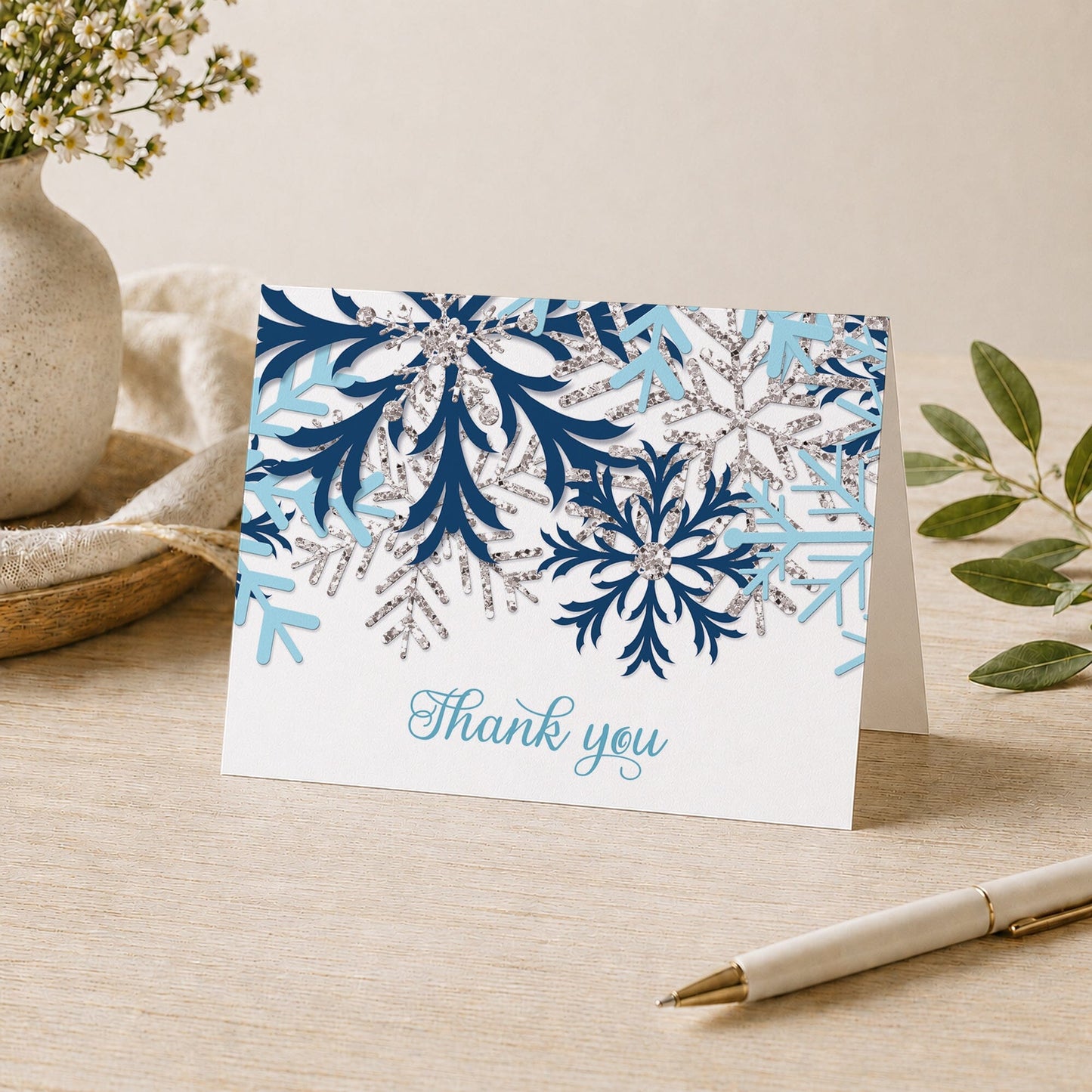 Winter Blue Silver Snowflake Thank You Card shown on a tabletop with a vase, plants, and a pen.