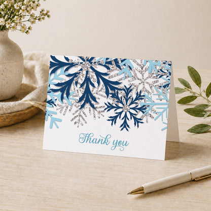 Winter Blue Silver Snowflake Thank You Card shown on a tabletop with a vase, plants, and a pen.