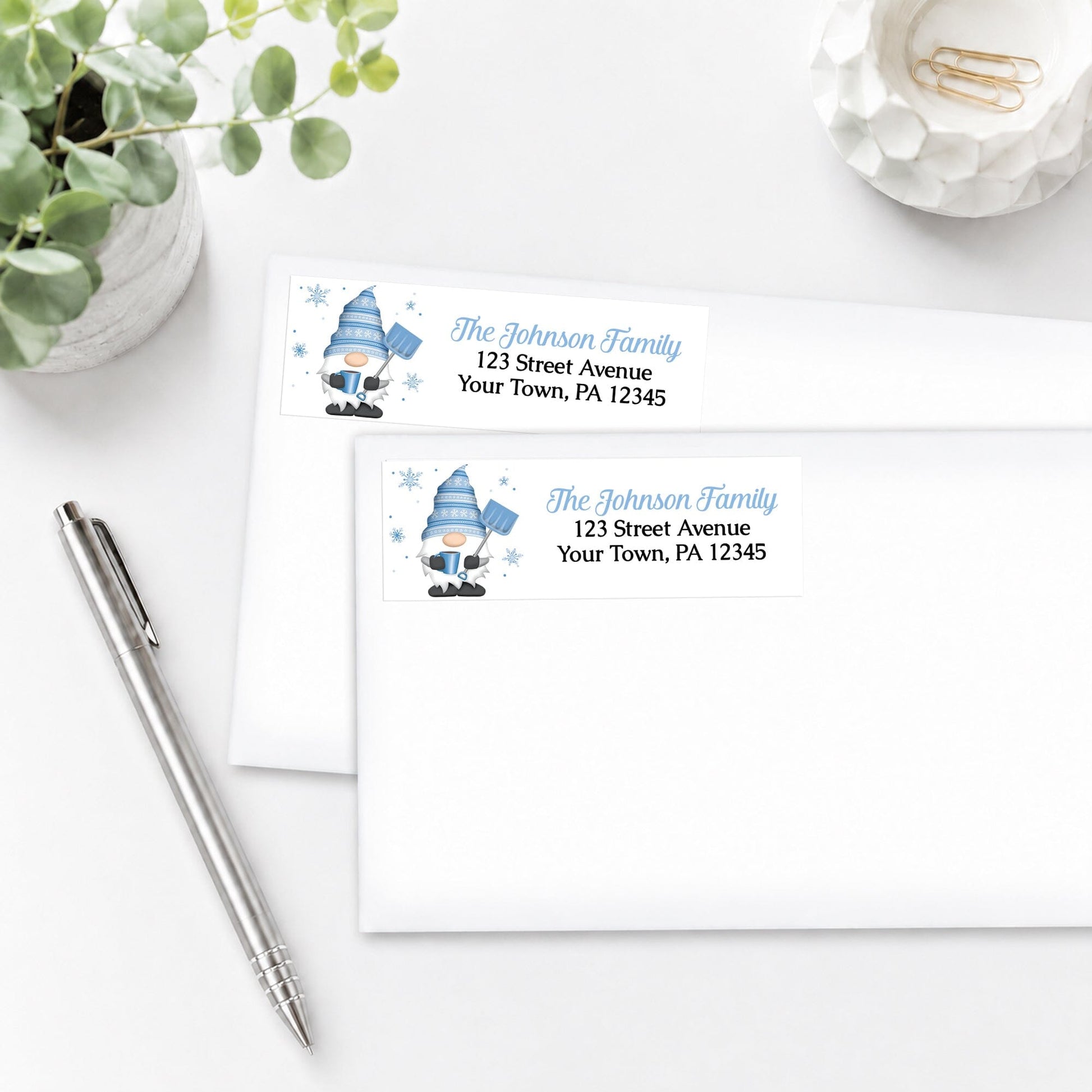 Two Winter Gnome Address Labels with a blue gnome design, shown on white envelopes, on a white surface with a pen and plant.