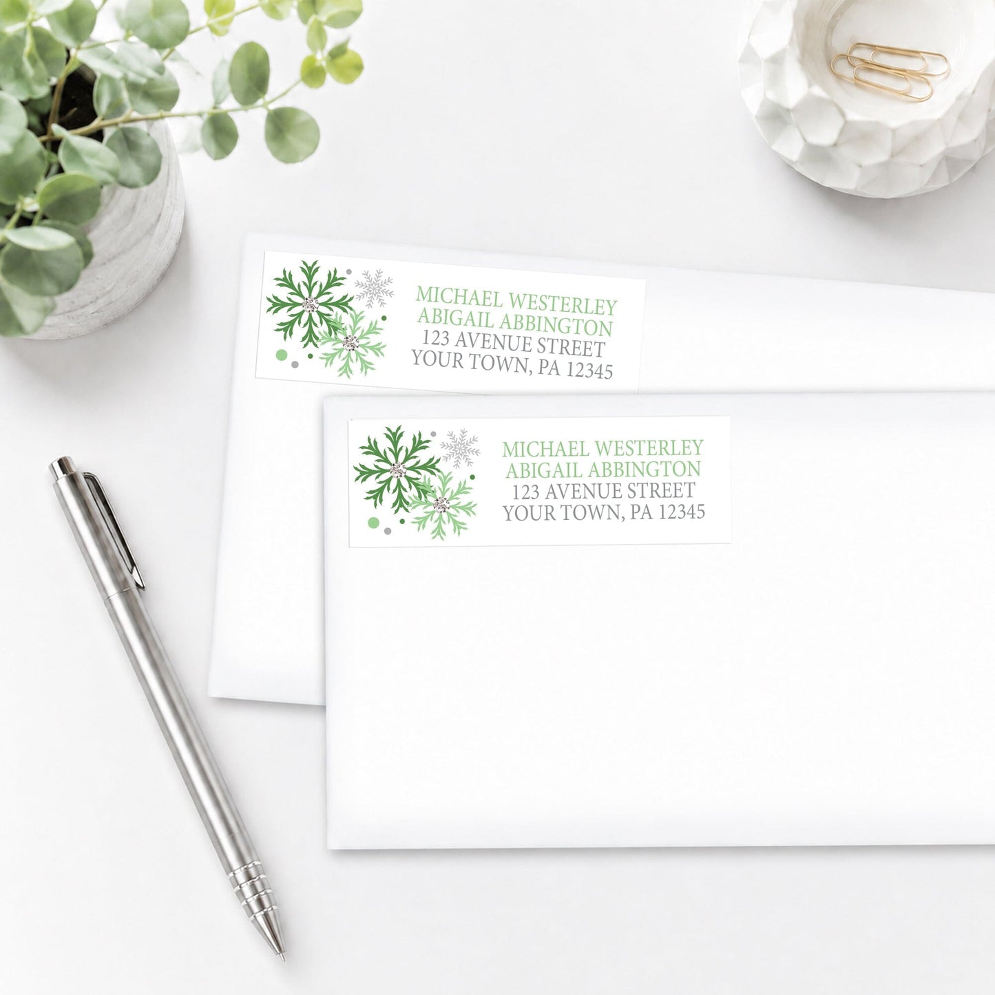 Winter Green Silver Snowflake Address Labels, shown on white envelopes, on a white surface with a plant and a pen.