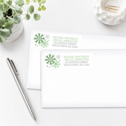 Winter Green Silver Snowflake Address Labels, shown on white envelopes, on a white surface with a plant and a pen.