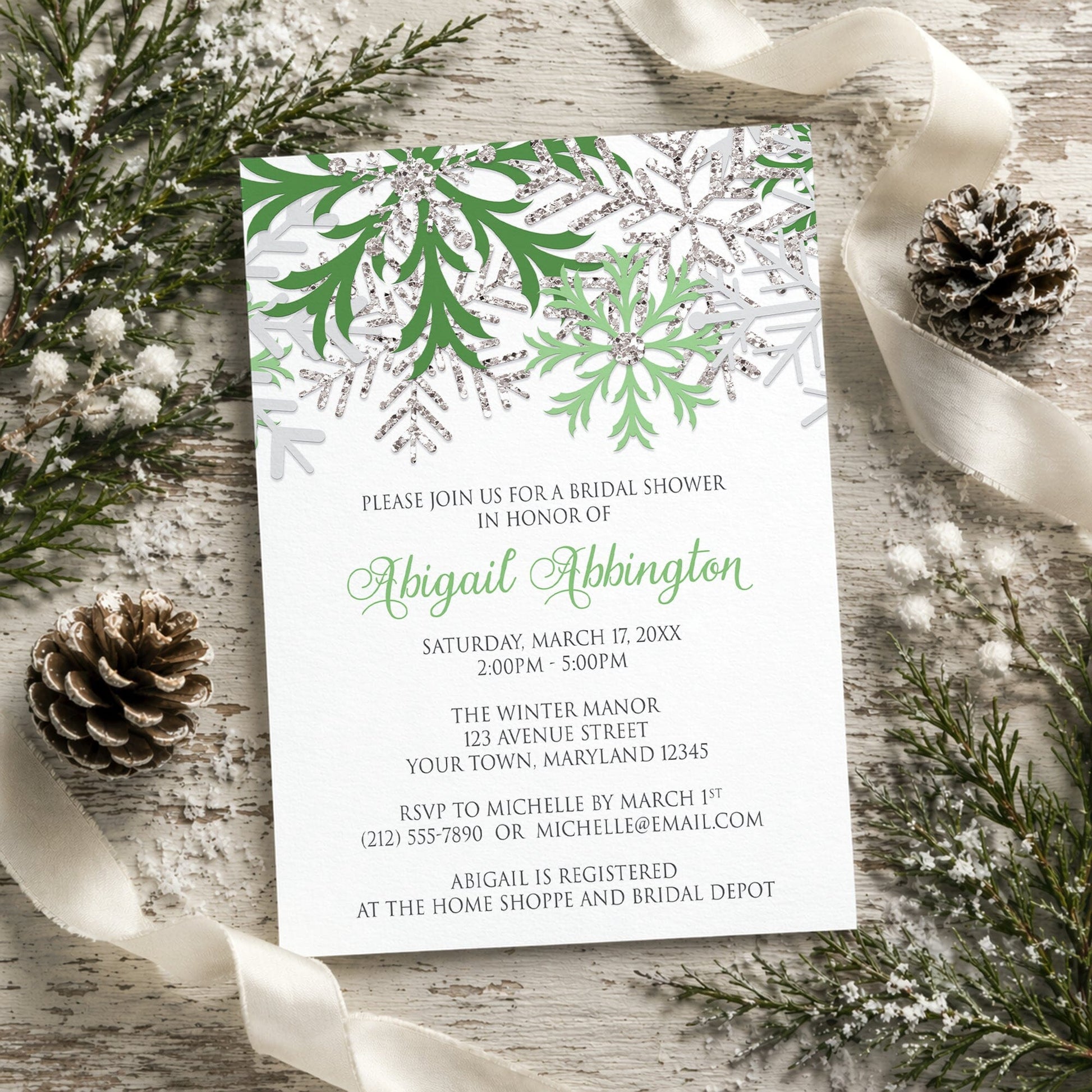 Winter Green Silver Snowflake Bridal Shower Invitation shown on a rustic surface with winter greenery, pine cones, and ribbons.