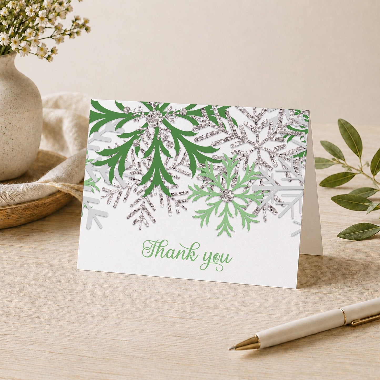 Winter Green Silver Snowflake Thank You Card shown on a desk with a vase, plants, and a pen.