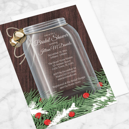Winter Mason Jar Pine Boughs Bridal Shower Invitation shown at an angle on a marble surface with a white envelope.