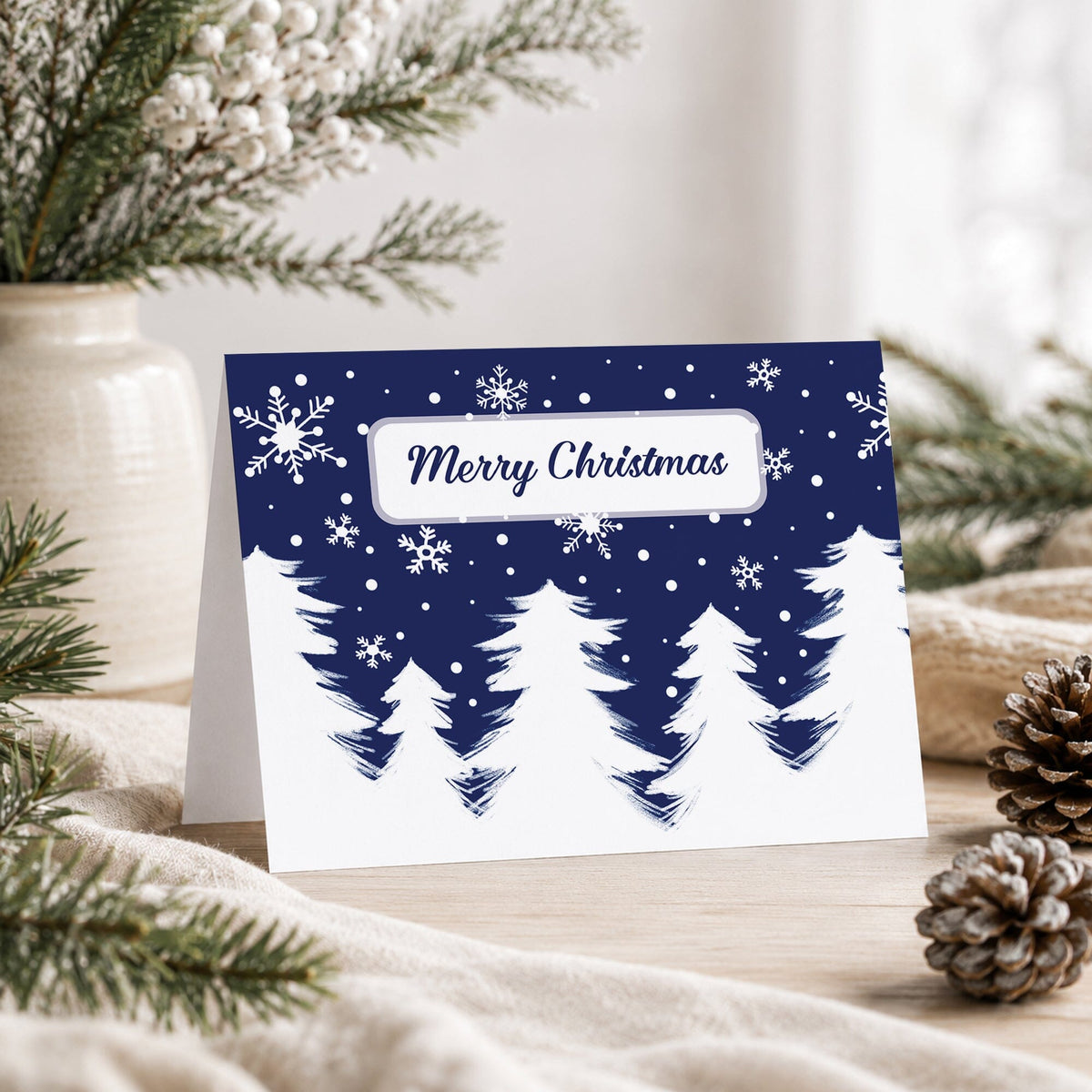 Winter Night Snow Christmas card with white snowy evergreen trees, falling snowflakes, dark navy blue sky, and Merry Christmas script inside a white frame, displayed on a wooden surface with pine branches, a vase, and a blanket in a styled indoor setting.