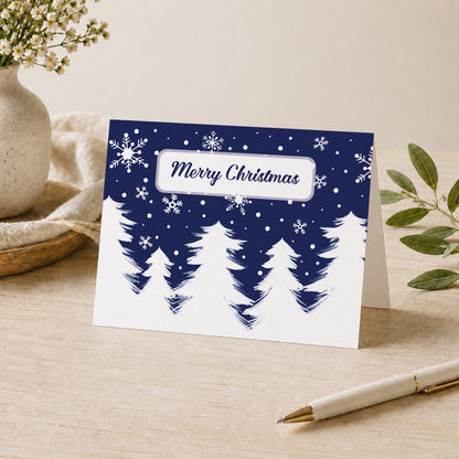 Winter Night Snow Christmas card with white snowy evergreen trees, falling snowflakes, dark navy blue sky, and Merry Christmas script inside a white frame, standing on a desk with a vase, plants, and a pen.