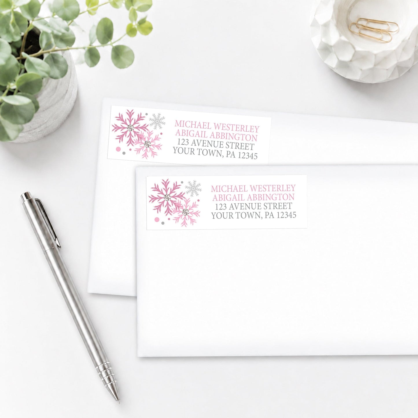 Address labels with pink snowflakes on white envelopes with a pen and plant in the background.