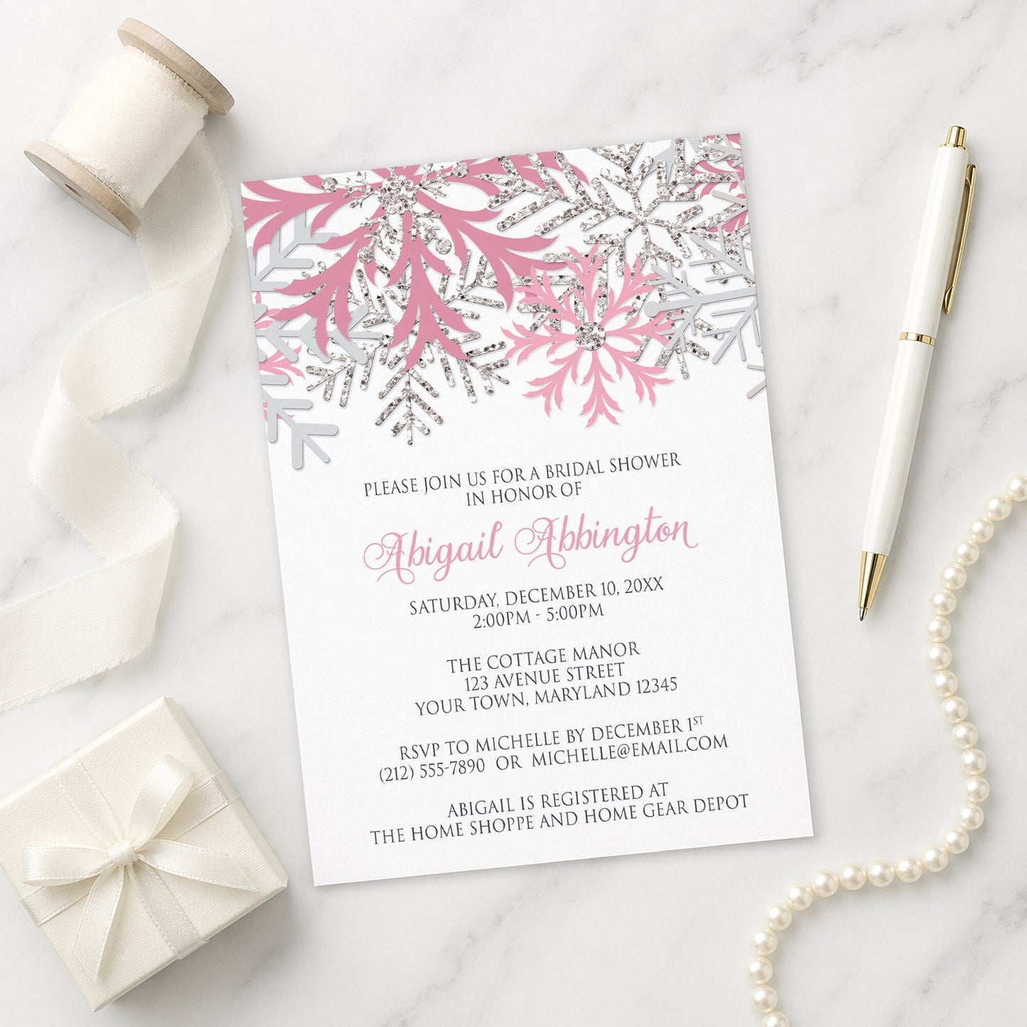 Winter bridal shower invitation with pink and silver snowflake design on a marble surface with gift, pen, and pearls.