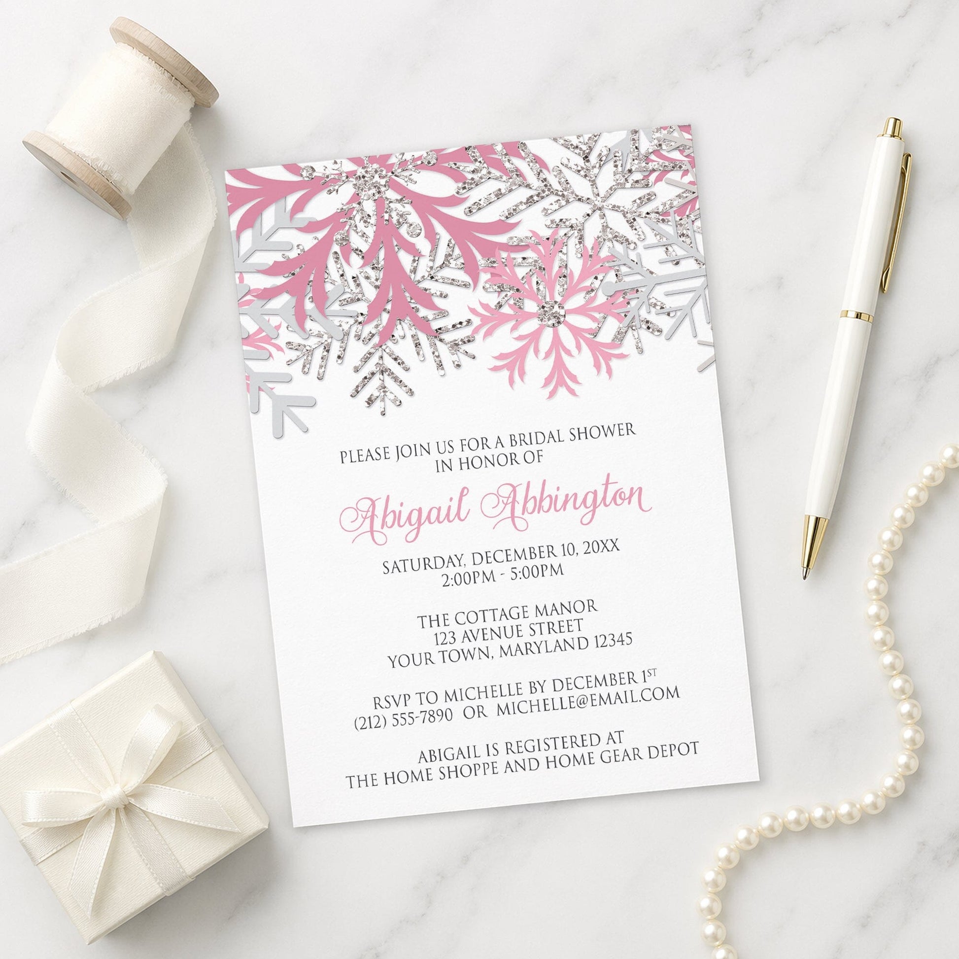Winter bridal shower invitation with pink and silver snowflake design on a marble surface with gift, pen, and pearls.
