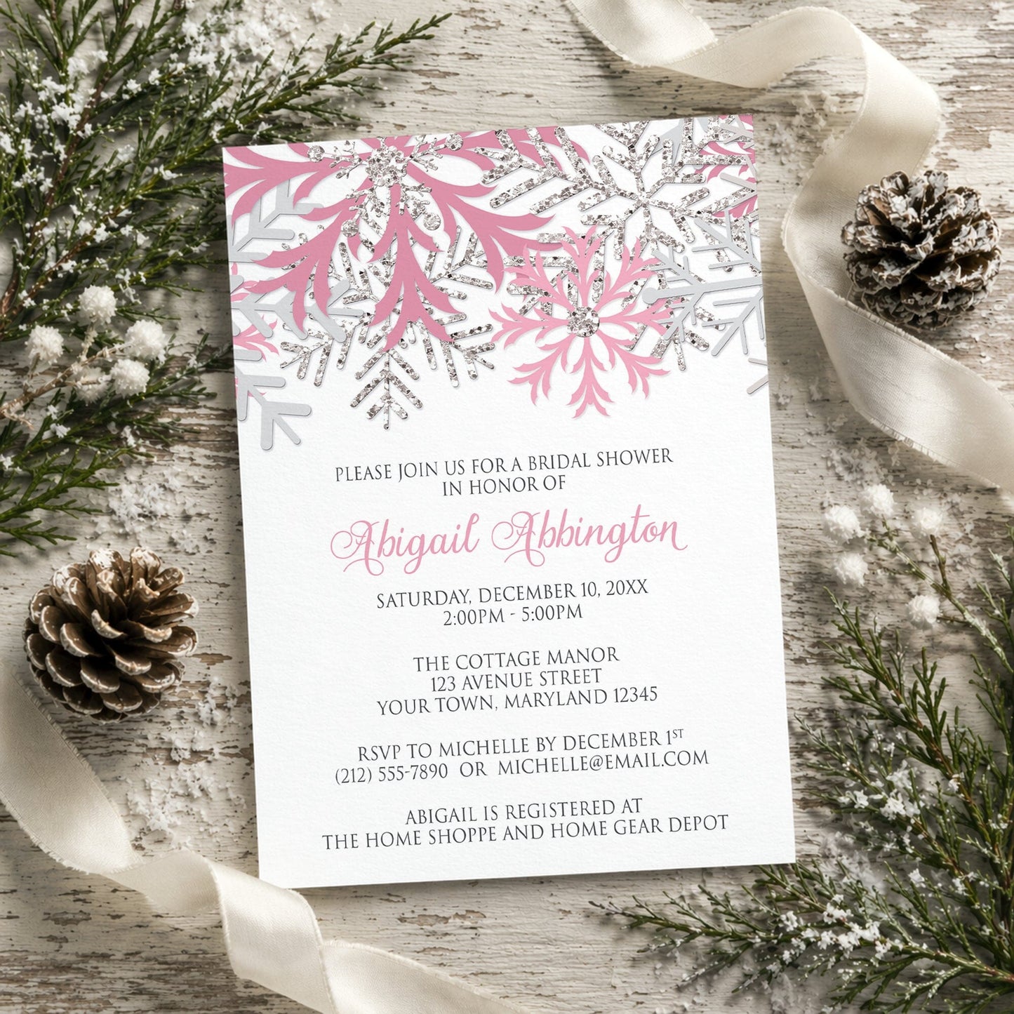 Winter bridal shower invitation with pink and silver snowflake design shown on a rustic wooden background with pine cones and ribbons.