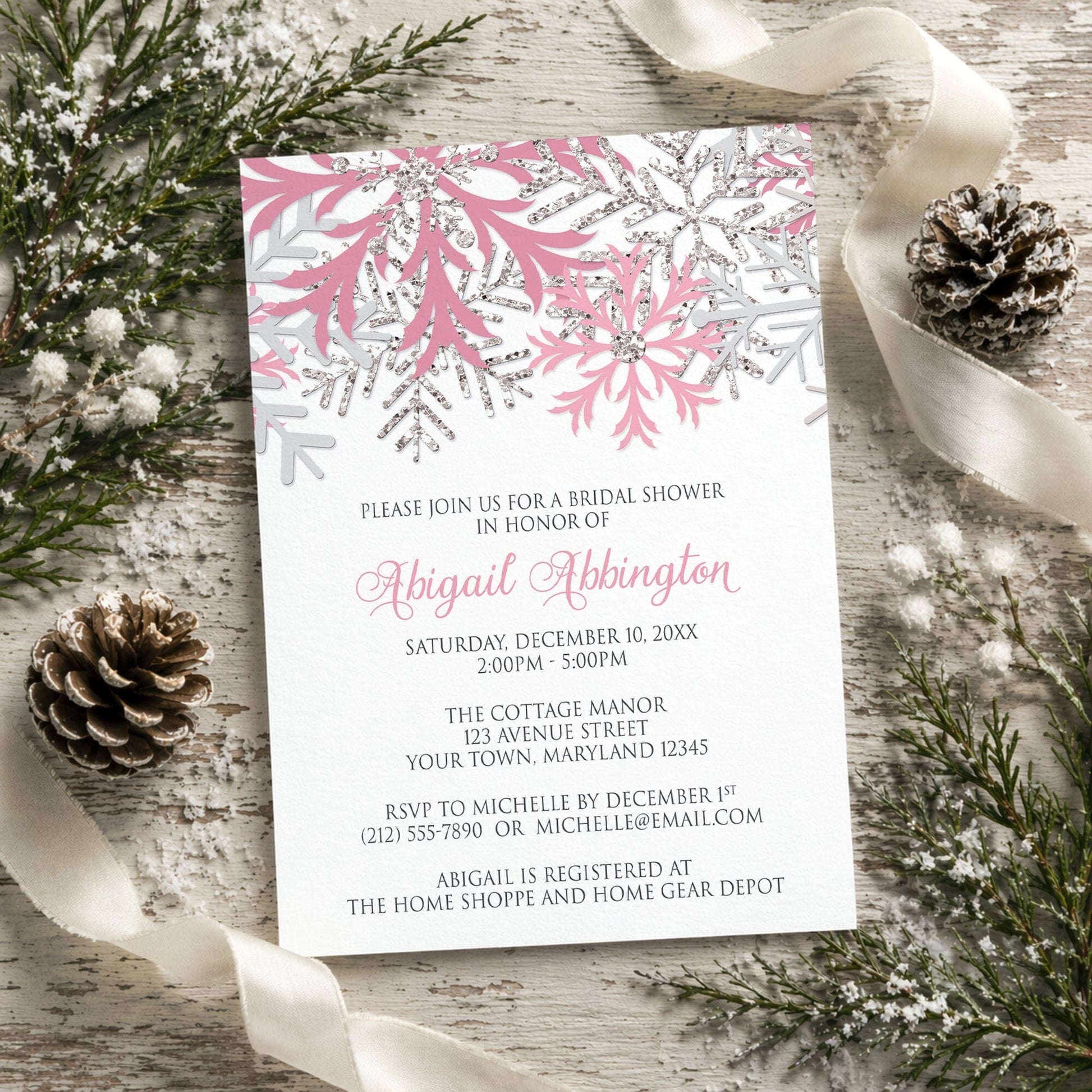 Winter bridal shower invitation with pink and silver snowflake design shown on a rustic wooden background with pine cones and ribbons.