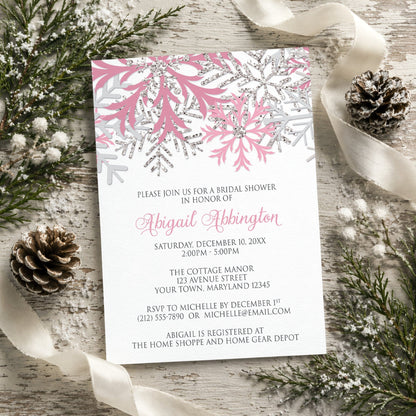 Winter bridal shower invitation with pink and silver snowflake design shown on a rustic wooden background with pine cones and ribbons.