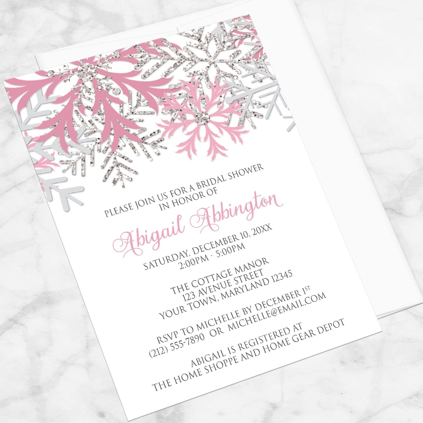 Winter bridal shower invitation with pink and silver snowflake design shown at an angle on a marble surface with a white envelope.