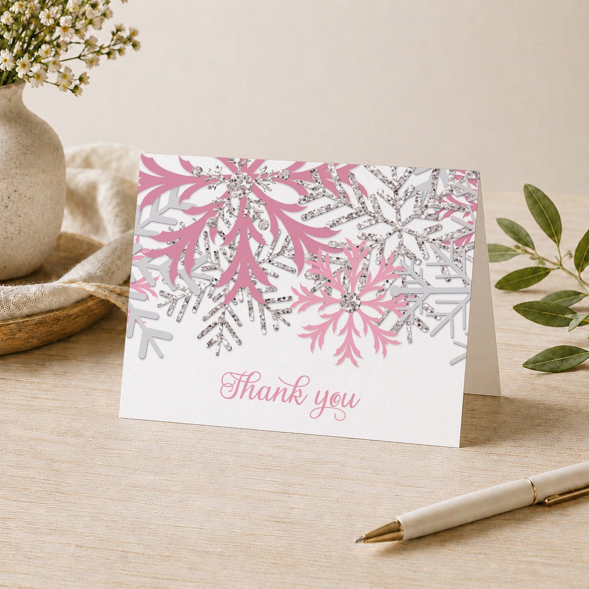 Winter Pink Silver Snowflake Thank You Card shown on a desk with a vase, plants, and a pen.