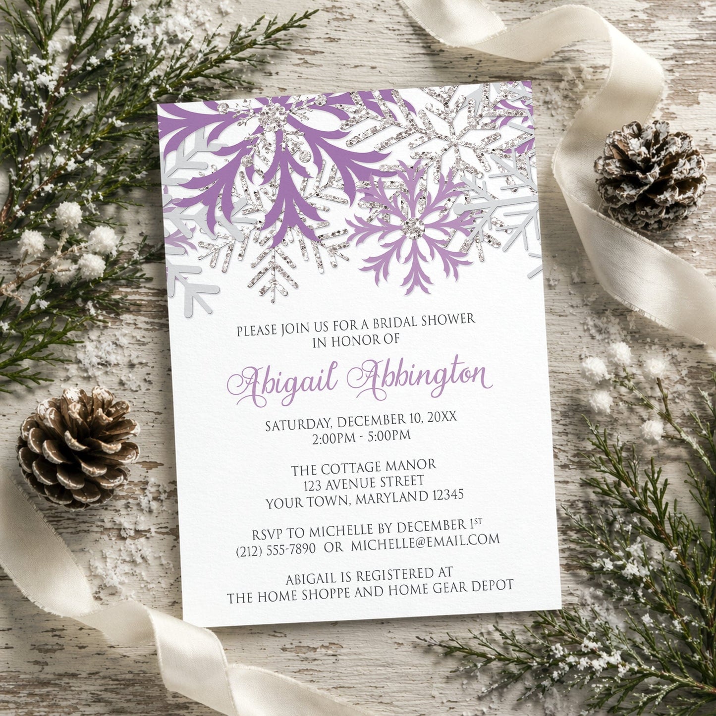 Winter bridal shower invitation with purple and silver snowflakes shown on a wooden surface with winter greenery, pine cones, and ribbon.