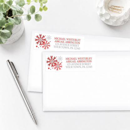 Two address labels with red snowflake designs on white envelopes shown on a white surface with a pen and plant.