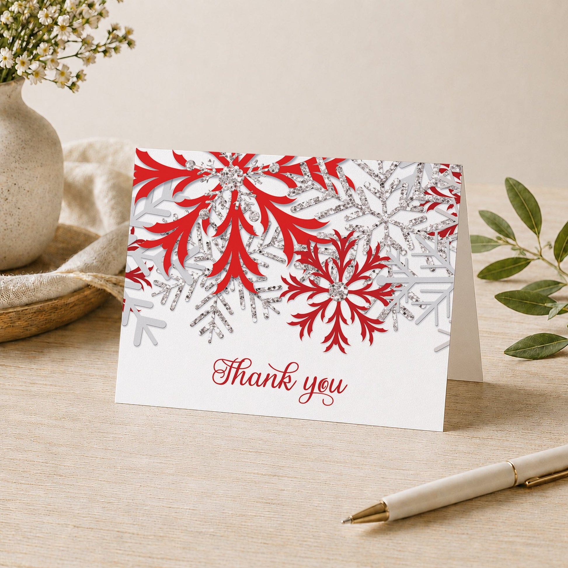 Winter Red Silver Snowflake Thank You Card shown on a desk with a vase, plants, and a pen.