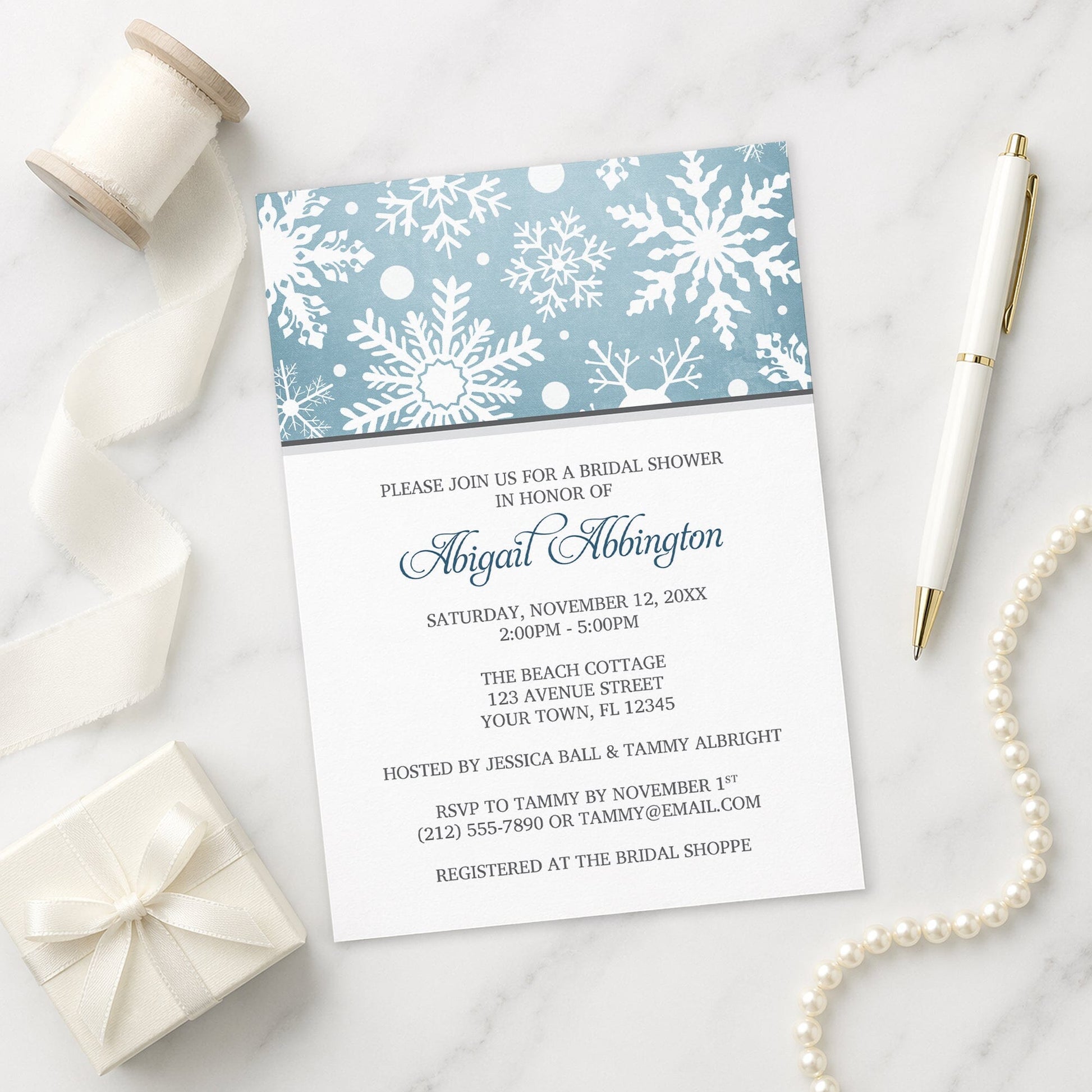 Winter Snowflake Blue Gray Bridal Shower Invitation shown on a marble surface with a white gift box, pen, ribbon, and pearls.