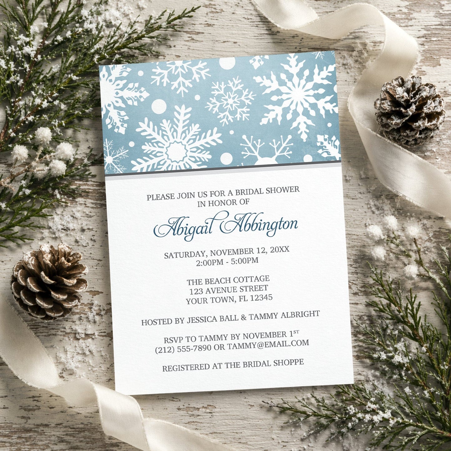 Winter Snowflake Blue Gray Bridal Shower Invitation shown on a rustic surface with winter greenery, pine cones, and ribbons.
