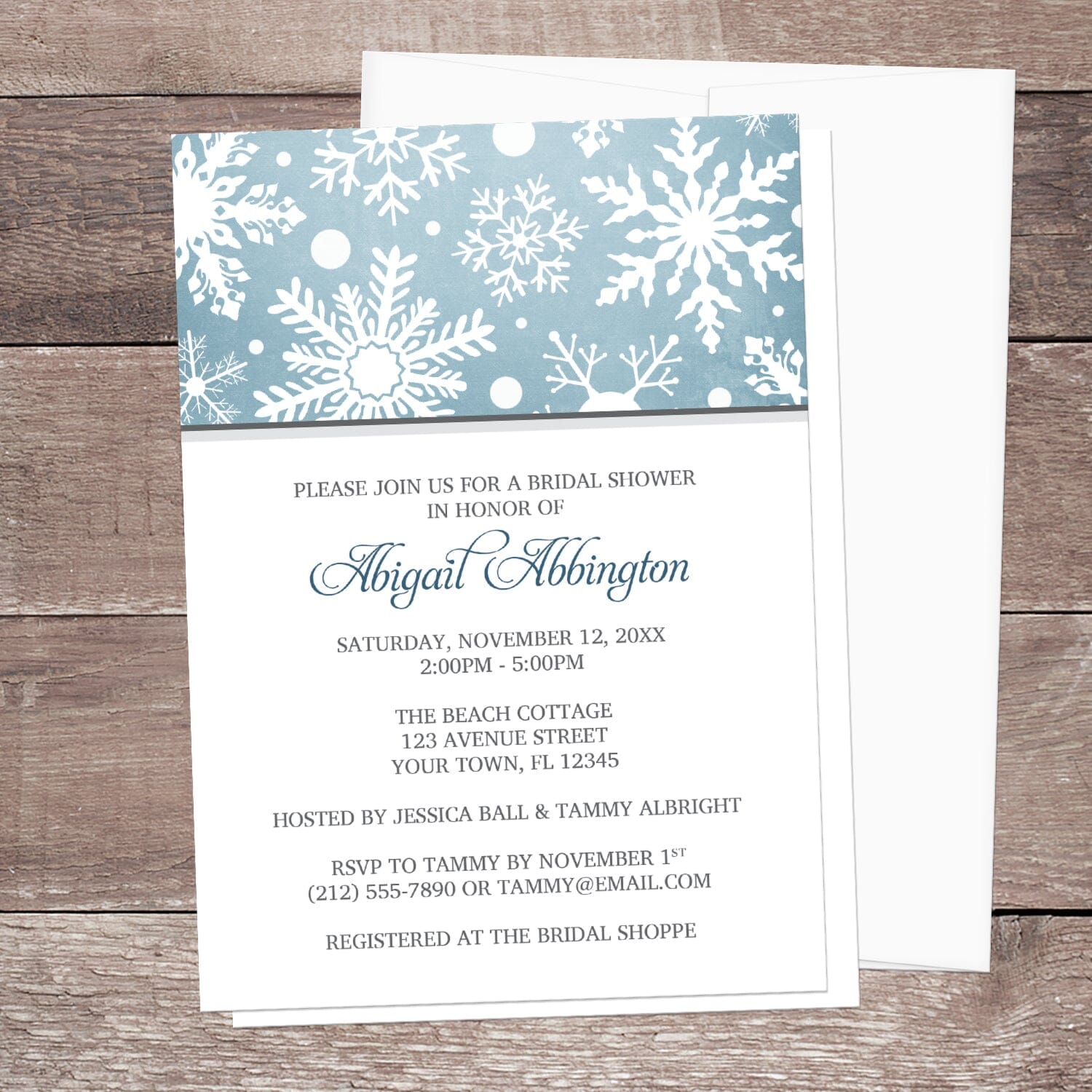 Winter Snowflake Blue Gray Bridal Shower Invitation shown on a wooden surface with a white envelope.