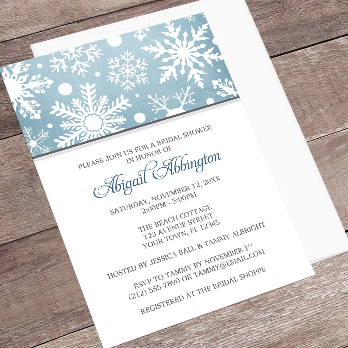 Winter Snowflake Blue Gray Bridal Shower Invitation shown at an angle on a wooden surface with a white envelope.
