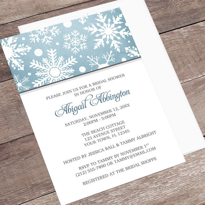 Winter Snowflake Blue Gray Bridal Shower Invitation shown at an angle on a wooden surface with a white envelope.