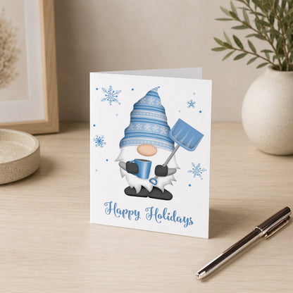 A white holiday greeting card featuring a blue and white gnome holding a snow shovel and mug, with snowflakes and the text 'Happy Holidays' on a light wood desk. A pen, framed art, and a potted plant are visible in the background.