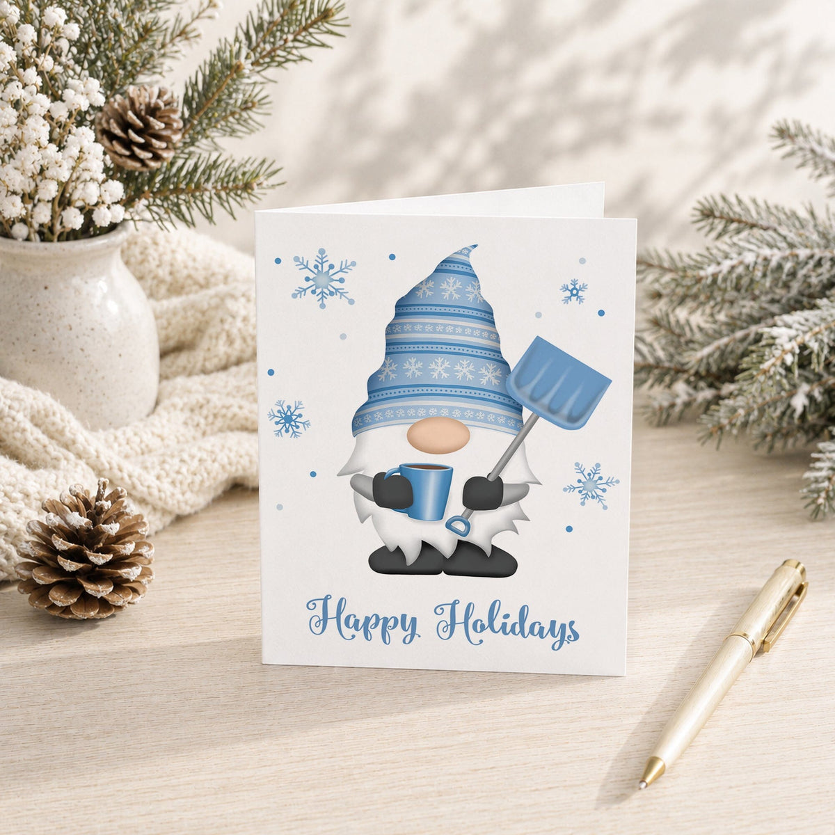 A white holiday greeting card featuring a blue-and-white gnome holding a snow shovel and mug, with snowflakes and the words 'Happy Holidays' printed below. The card is styled on a light wood surface with pine branches, a cream knit blanket, a vase, and a gold pen.