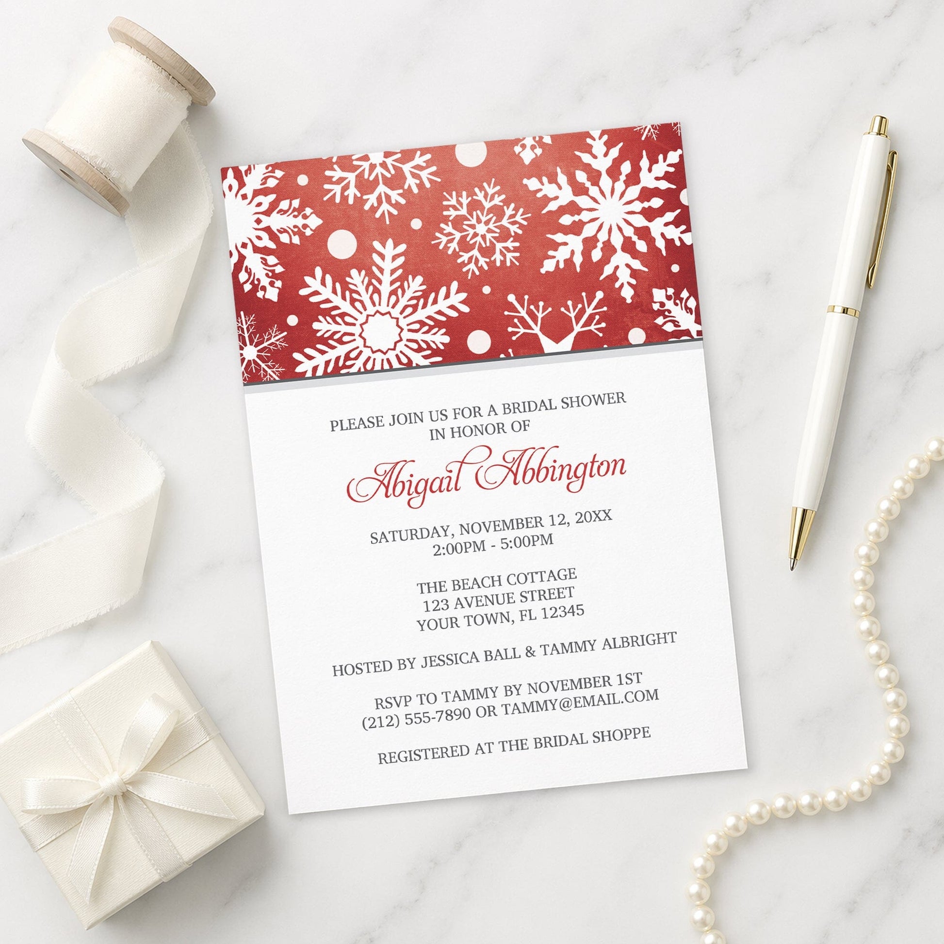 Winter Snowflake Red Gray Bridal Shower Invitation shown on a marble surface with a white gift box, pen, ribbon, and pearls.