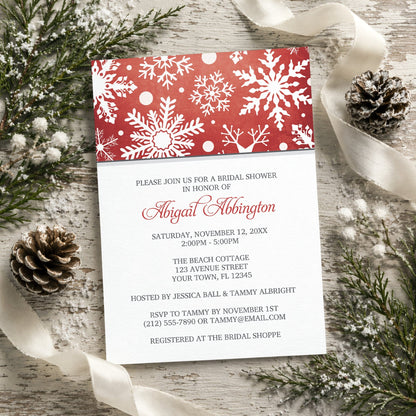 Winter Snowflake Red Gray Bridal Shower Invitation shown on a rustic surface with winter greenery, pine cones, and ribbons.