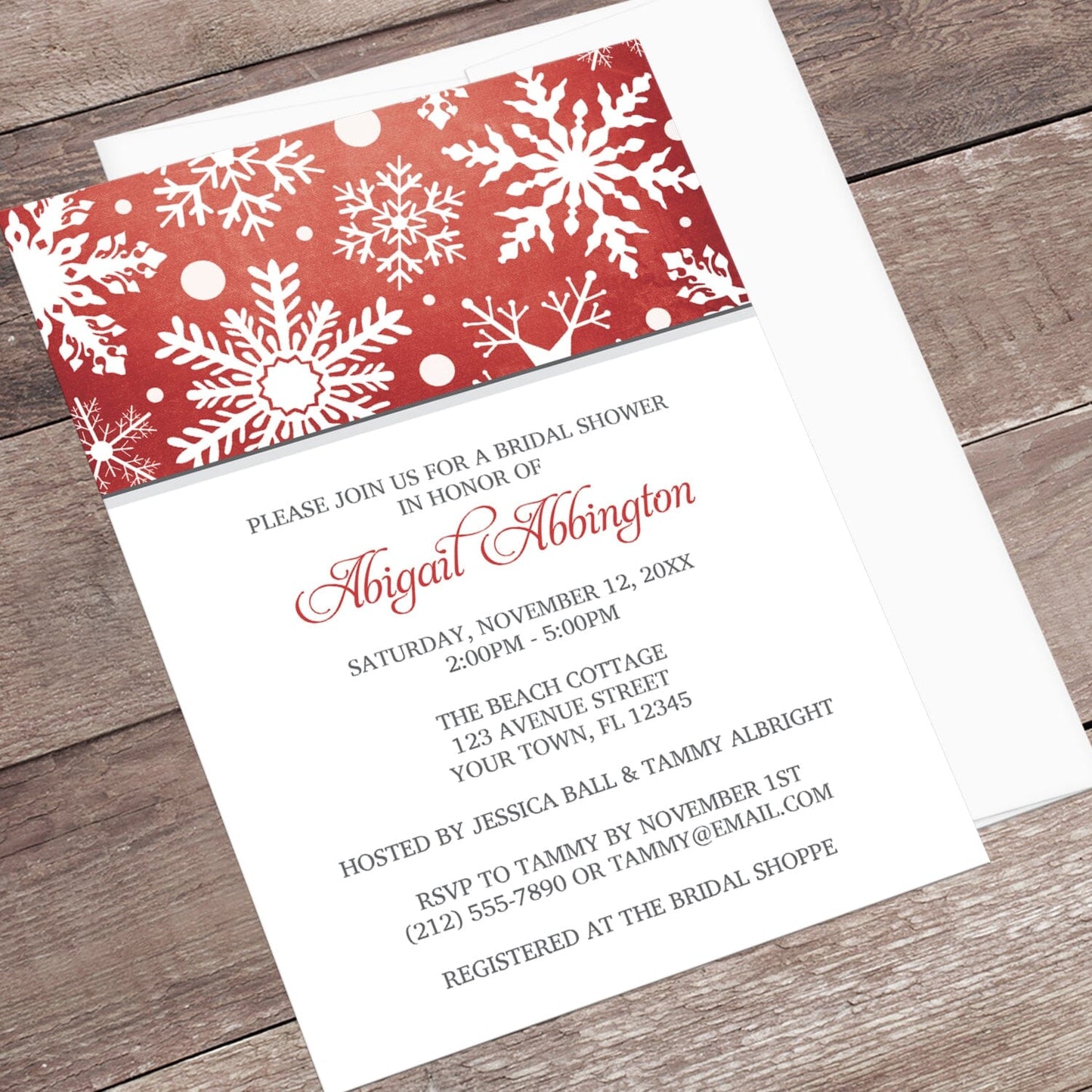 Winter Snowflake Red Gray Bridal Shower Invitation shown at an angle on a wooden surface with a white envelope.