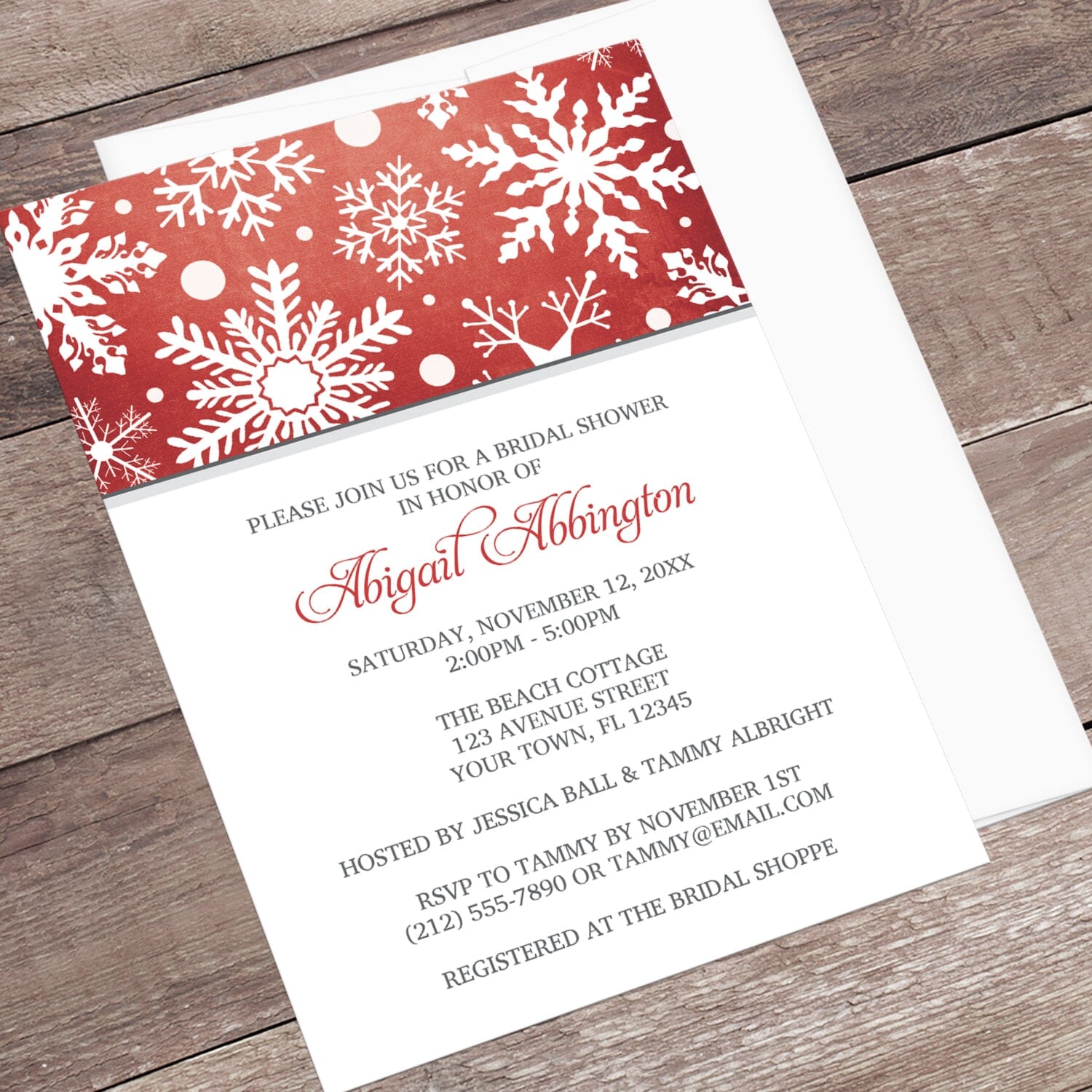 Winter Snowflake Red Gray Bridal Shower Invitation shown at an angle on a wooden surface with a white envelope.