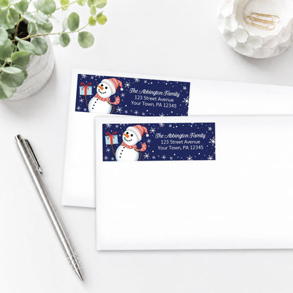 Winter Snowman Address Labels on white envelopes, shown on a white surface with a plant and a pen.