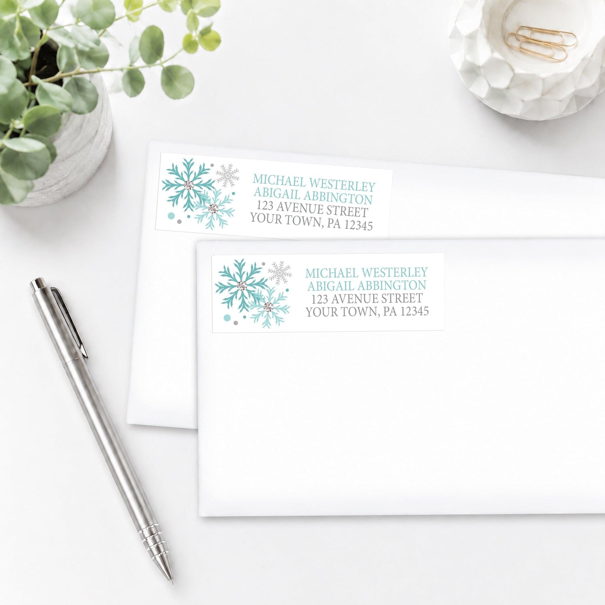 Winter Teal Silver Snowflake Address Labels on white envelopes on a white surface with a pen and plant.