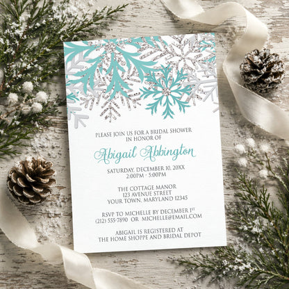 Winter Teal Silver Snowflake Bridal Shower Invitation shown on a rustic wood surface with winter greenery, pine cones, and ribbons.