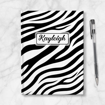 Personalized Zebra Print Journal at Artistically Invited. A custom book illustrated with a bold and sleek black and white zebra print pattern that wraps around the cover of the journal. Your personalized name is custom printed in a cute script font in black within a white rectangular design on the front of the book over the zebra print background. Image shows the journal next to a pen.